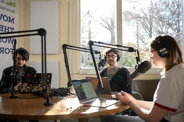three students sit in front of microphones in the podcast studio