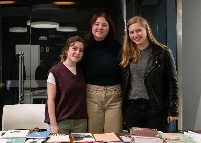 在奥尔登图书馆，三个学生在一张桌子后面摆姿势拍照，桌子上放着书.